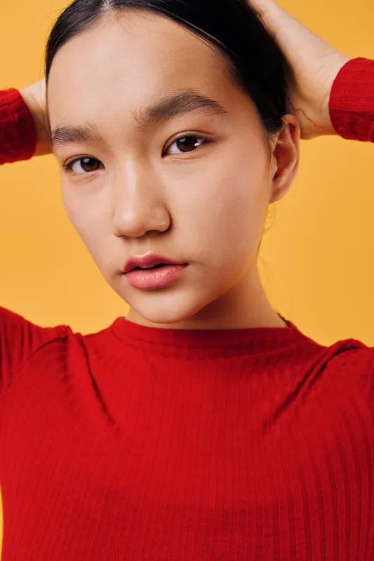 Young woman with dark hair in red shirt poses with hands behind head on orange background, showing natural skin and neutral expression in closeup portrait photography.