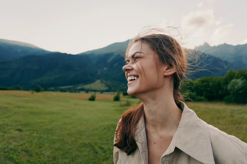 Bright woman enjoys outdoor life in a sunlit field with distant mountains, candid smile, casual outfit, relaxed mood, lifestyle portrait captured in nature
