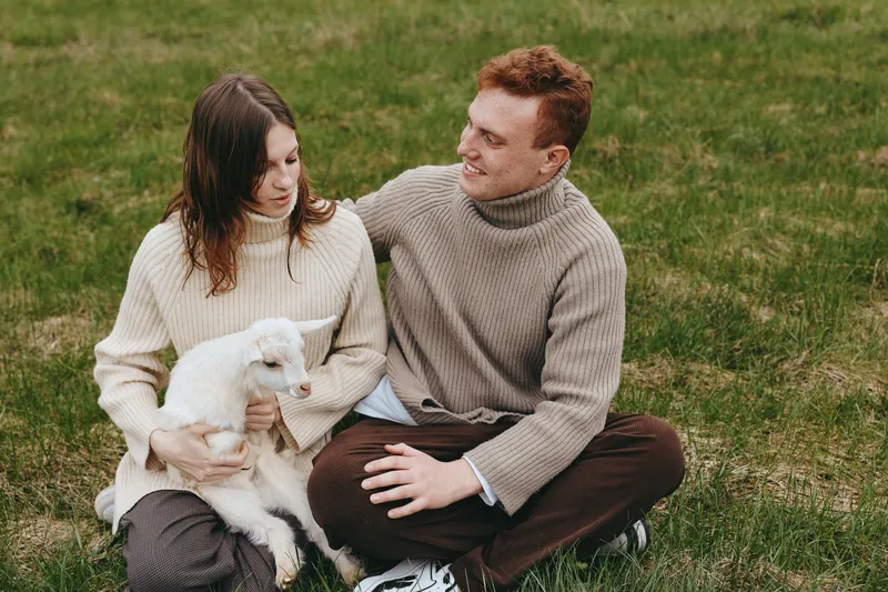 A relaxed couple sits on grass with a small white dog, sharing a tender moment in a sunny afternoon. A lifestyle portrait of companionship, warmth, and everyday connection.