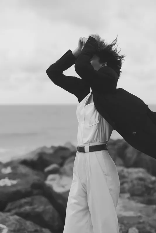 Wind sweeps across a rocky shore, revealing a stylish figure in white and dark coat, creating dynamic fashion and freedom in a moody, cinematic outdoor moment