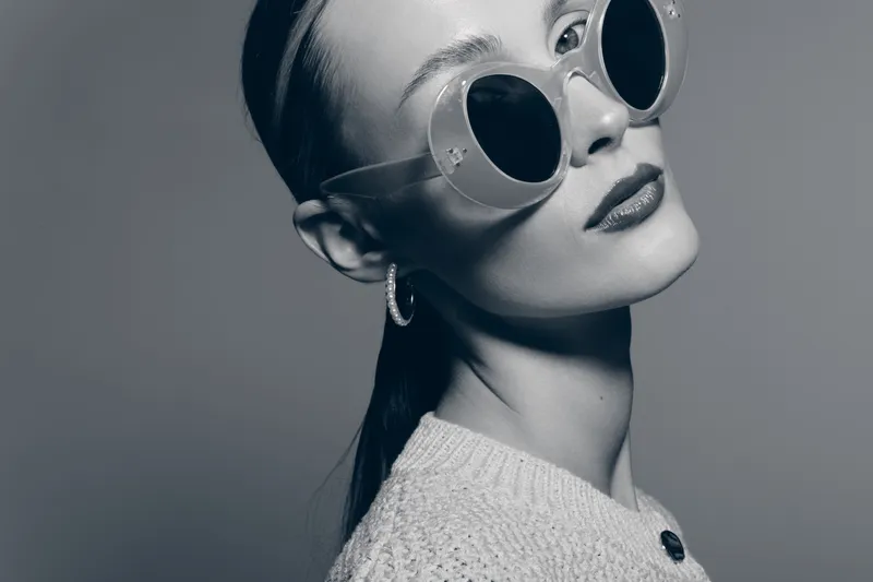 Stylish white woman with pale skin and slicked hair wearing round sunglasses and knitted sweater isolated on grey.