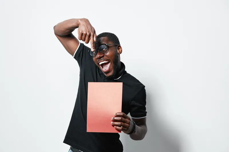African American man with dark skin and glasses holding blank paper and pointing at it with excitement in studio. Casual clothing and joyful expression on white background.