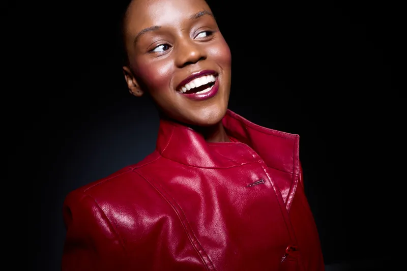 Smiling dark skinned African woman wearing a shiny red leather jacket looks to the side with joy. Studio portrait on black background. Concept of fashion, beauty, confidence, and style.