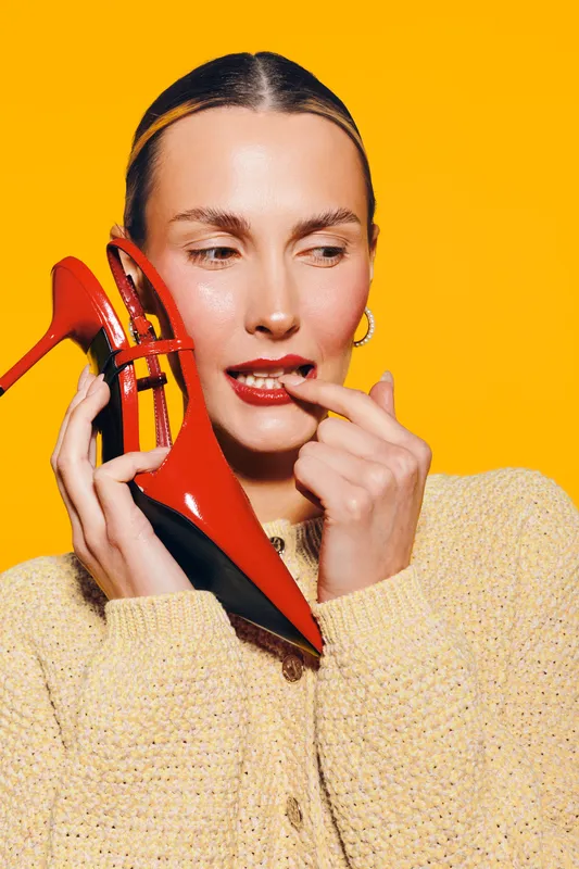 Young woman in beige sweater holds red high heel shoe near face, biting finger. Bright yellow background creates vibrant contrast. Concept of fashion, style, and playful mood.