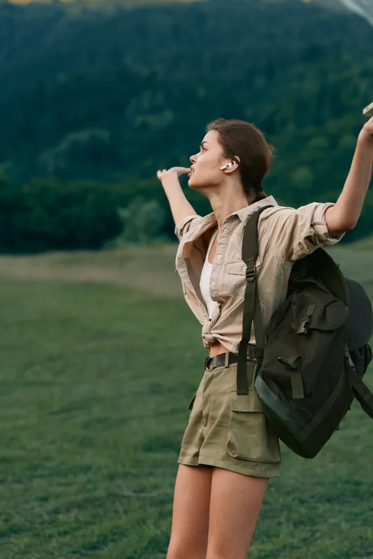 Adventurous woman in khaki shorts and tan shirt raises her arm in a green field, backpack on, expressing freedom and celebration on a sunny day with distant hills.
