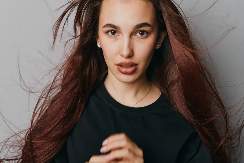 Young woman with long hair wearing black shirt looking at camera with serious expression on gray background. Portrait of female model with natural makeup and loose hair.