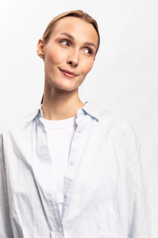 Young woman with light skin and blonde hair looks to the side with a subtle smile. She wears a white shirt and a light blue shirt over it against a plain white background.