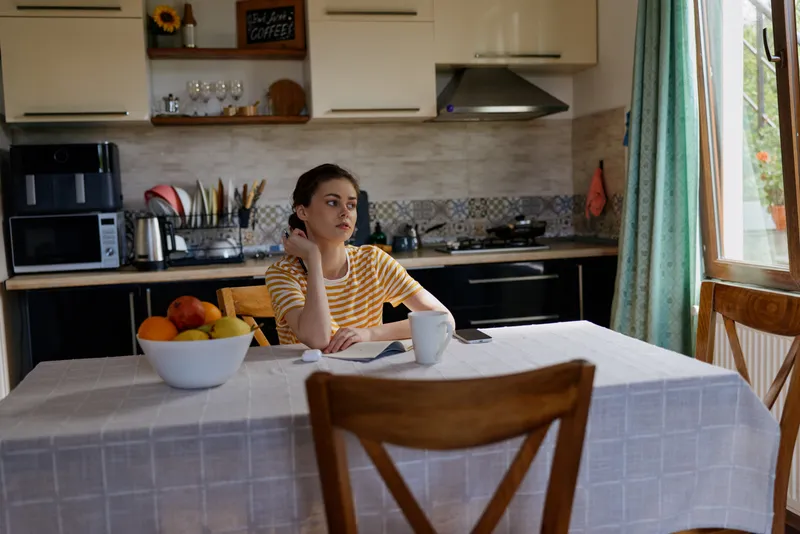 A pensive woman sits at a bright kitchen table, notebook in hand and a mug beside her, while a bowl of fruit appears in the foreground during a quiet morning routine.