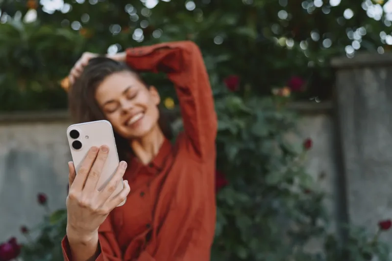 A cheerful woman in a red top captures a selfie in a sunny garden, posing with hair loose and a bright smile. Casual lifestyle moment focused on self expression and outdoor joy