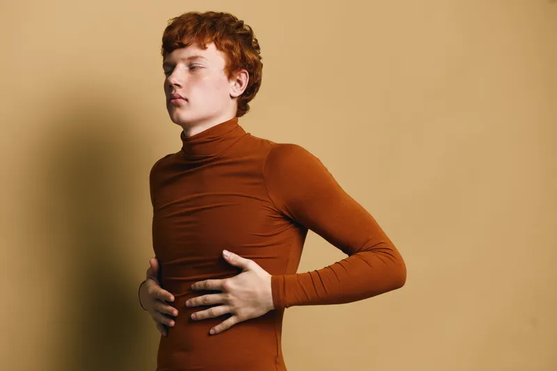 Young man with red hair and closed eyes wearing tight brown turtleneck standing against plain beige background with hands on stomach in thoughtful pose.