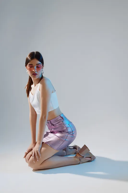 Young woman in a white top and shiny pink skirt kneels on the floor wearing stylish sunglasses. Studio shot with soft lighting. Concept of woman beauty fashion and modern style.