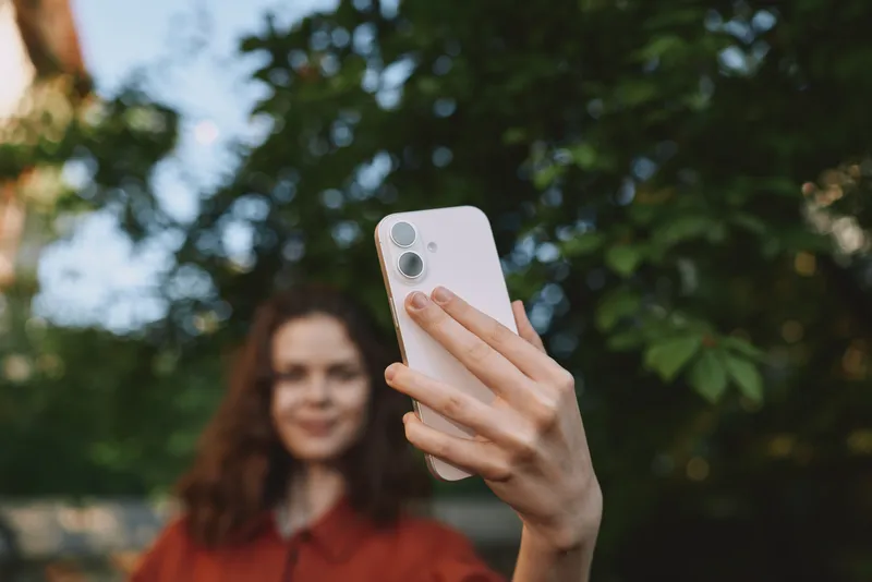 Smiling woman outdoors capturing a selfie with a smartphone, casual attire, lush garden backdrop, candid lifestyle moment, warm natural light and relaxed mood, in frame, vibrant
