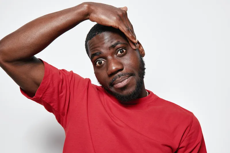 African man with dark skin wearing red t shirt touching forehead with hand looking confused or stressed isolated on white background. Male portrait with short hair and beard expression.
