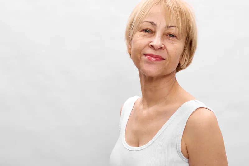 Older blonde woman smiling in studio portrait, white sleeveless top, calm face closeup, soft lighting, neutral background, copy space