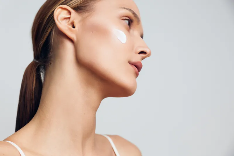 Young woman with smooth skin applies cream on cheek looking sideways showing natural beauty and skincare routine isolated on light background with soft lighting and calm expression.