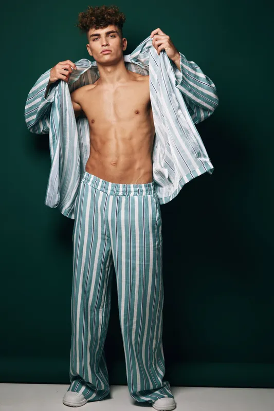 Young man with curly hair wearing striped pajamas and white sneakers standing against dark background showing open shirt and toned abs with confident expression.