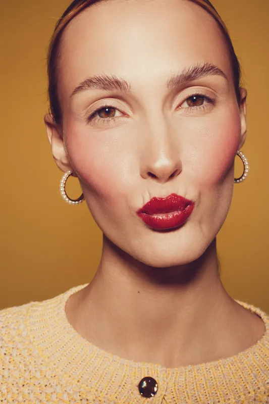 Close-up of a woman with red lipstick making a kiss face, wearing hoop earrings and a knitted sweater. Warm brown background highlights her smooth skin and confident expression.