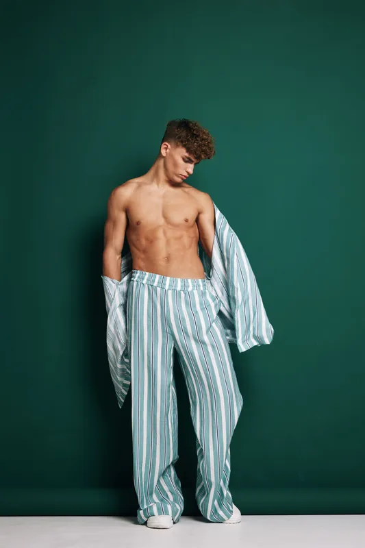 Young man with curly hair wearing striped loose pants and matching shirt open, standing against green background with bare chest and white sneakers in studio.
