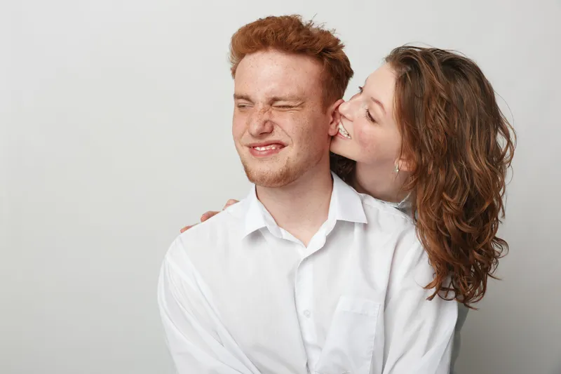 Young couple in white shirts share a playful moment, woman kissing man on cheek. Studio portrait with light background, concept of love, affection, and happiness.