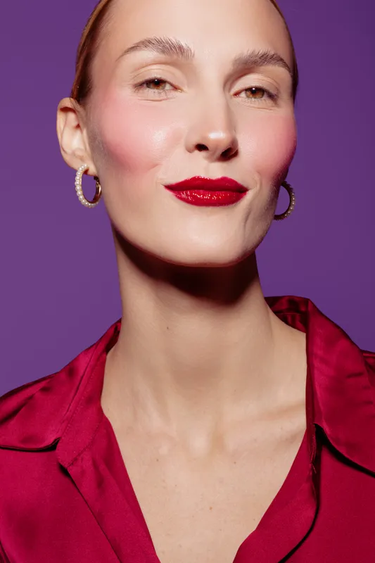 Confident woman with red lipstick and hoop earrings poses against purple background. She wears a red blouse and smiles slightly, showcasing beauty and fashion concepts.