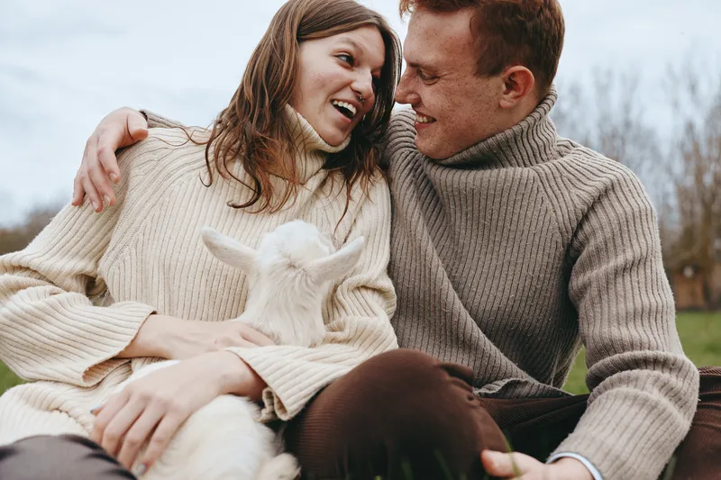 A smiling couple outdoors cuddle a curious goat kid, sharing a warm moment in soft sweaters. A candid lifestyle scene highlighting companionship, nature, and gentle animal interaction.