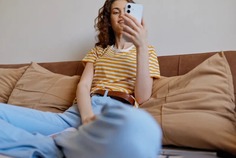 Young woman with curly hair sits on a beige sofa wearing a yellow striped tshirt and light jeans, taking a casual selfie in a cozy home living room with soft natural light and a relaxed mood.