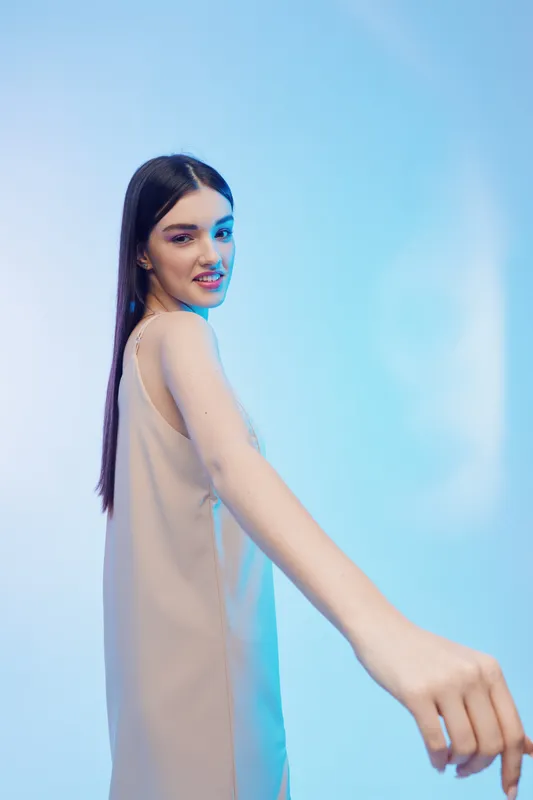 Young woman with long dark hair in a beige dress poses confidently against a blue background. Concept of beauty, fashion, and elegance in studio lighting.