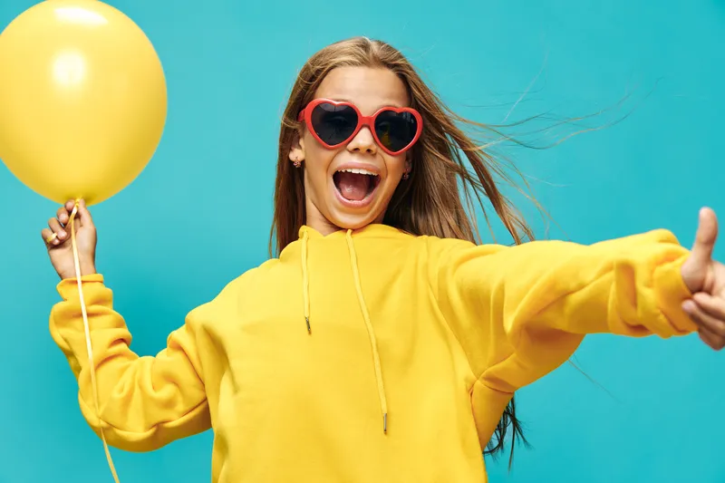 Girl wearing yellow hoodie and heart shaped sunglasses holding yellow balloon and showing thumbs up, smiling happily against blue background in casual style and joyful mood