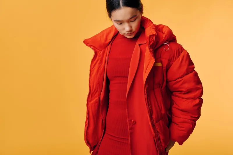 Woman wearing red coat and dress standing with head down against orange background. Young female model in fashion outfit with casual style and loose hair looking down.