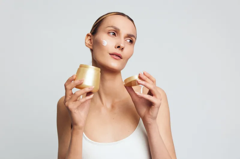 Young woman with fair skin applies cream on her face while holding an open jar of moisturizer against a plain light background in a skincare routine.