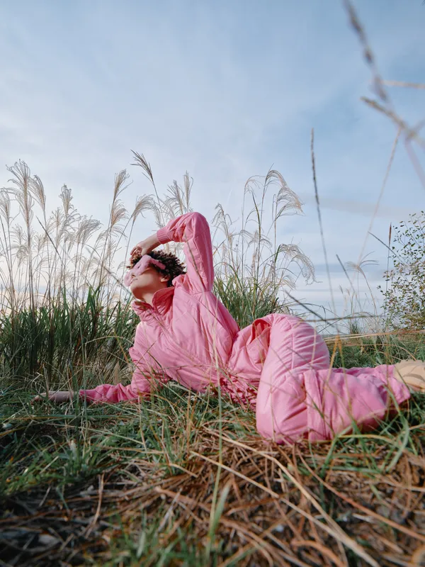 Pink jumpsuit worn by a woman resting on grass in an open meadow, wind in her hair, sunlit sky above for a serene outdoor fashion moment in a natural landscape.