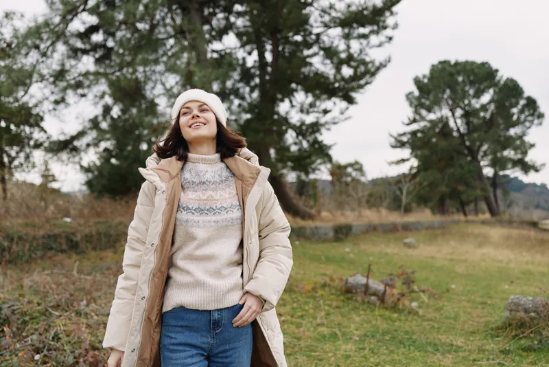 Woman in warm beige coat and beanie enjoys a breezy countryside day, smiling while strolling through a grassy field with scattered trees and soft overcast sky.