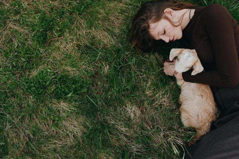 Young woman lies on grass holding a baby goat gently in her arms enjoying a peaceful moment outdoors in casual clothing with natural light and soft green background