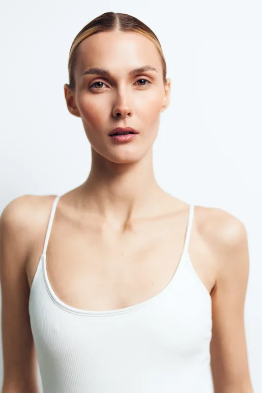 Woman portrait in studio closeup, calm beauty skincare concept, minimal white tank top, clean light background, neutral expression, copy space for wellness