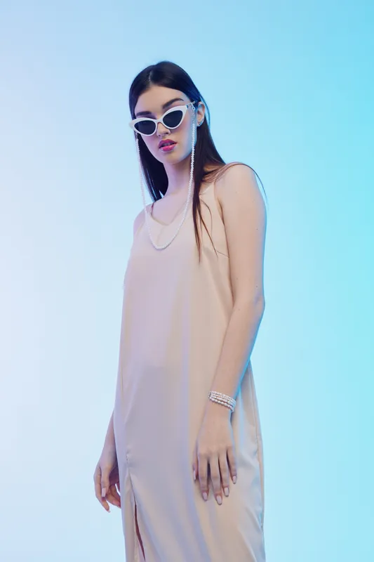 Young woman in a beige dress and white sunglasses poses against a blue background. Fashion and beauty concept emphasizing style and confidence in studio lighting.