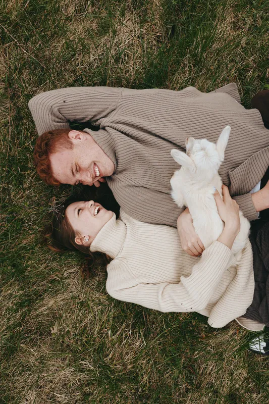 Happy couple relaxing on grass with a gentle goat kid, sharing a warm cuddle in a cozy outdoor moment, natural light and candid lifestyle scene, romantic and peaceful