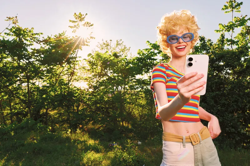 Young woman with curly blonde hair takes a playful selfie in bright sunlight wearing a striped crop top and blue sunglasses