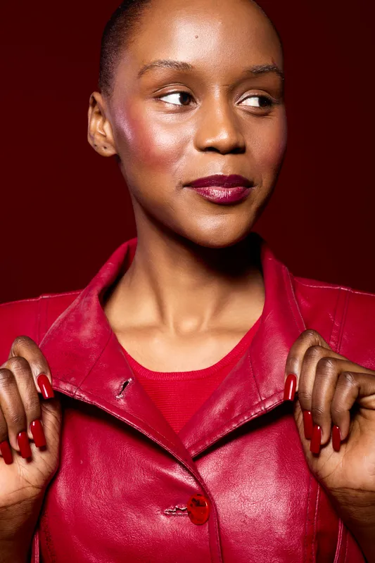 Portrait of confident black woman with dark skin wearing red leather jacket and matching nail polish. She looks to the side with a slight smile on red background. Concept of fashion and beauty.