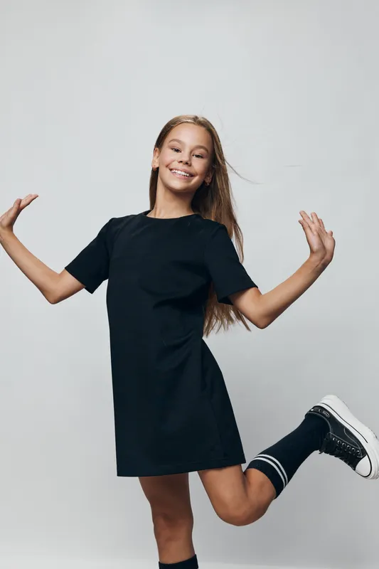 Teen girl in black dress and sneakers smiling joyfully while posing with one leg raised and arms outstretched on grey background for youth and happiness concept.