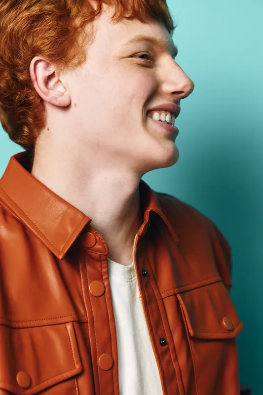 Young man with red hair smiling and looking sideways wearing brown leather jacket and white shirt on blue background, casual style and happy expression portrait.