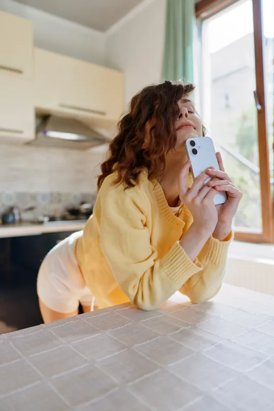 A woman in a yellow cardigan leans over a kitchen table, glancing up from her phone with a thoughtful gaze. A cozy home lifestyle moment, conveying contemplation and quiet daily life.