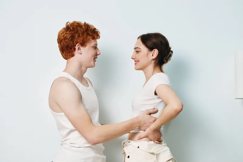 Young couple with casual clothes smiling and looking at each other in bright studio. Man and woman share affectionate moment with gentle touch and happy expressions.