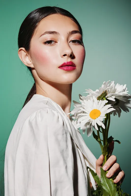 Portrait of a young woman with glossy dark hair and vibrant makeup holding white daisies. Clean fashion beauty shot on muted turquoise background. Concept of natural elegance and skincare.