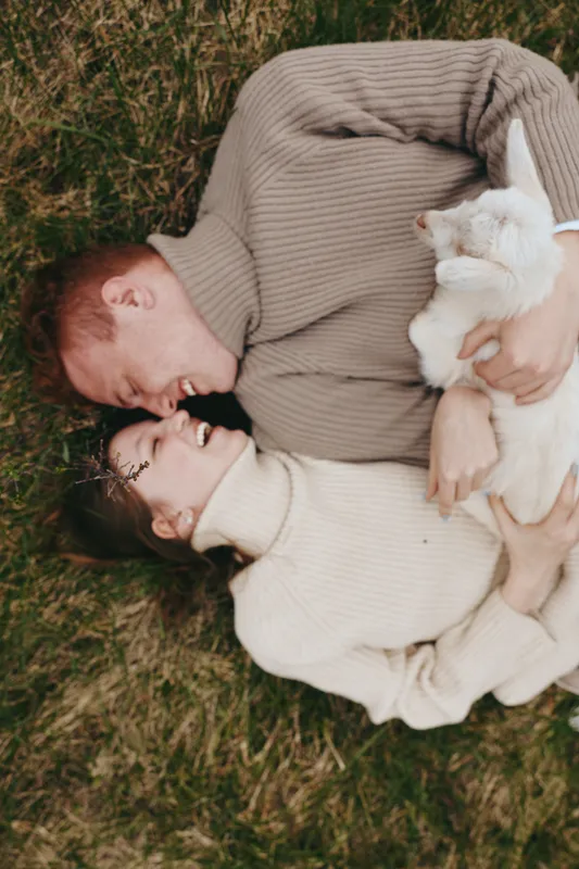 A young couple lies on the grass holding a white baby goat between them while smiling and enjoying a cozy lifestyle moment outdoors in knitted sweaters.