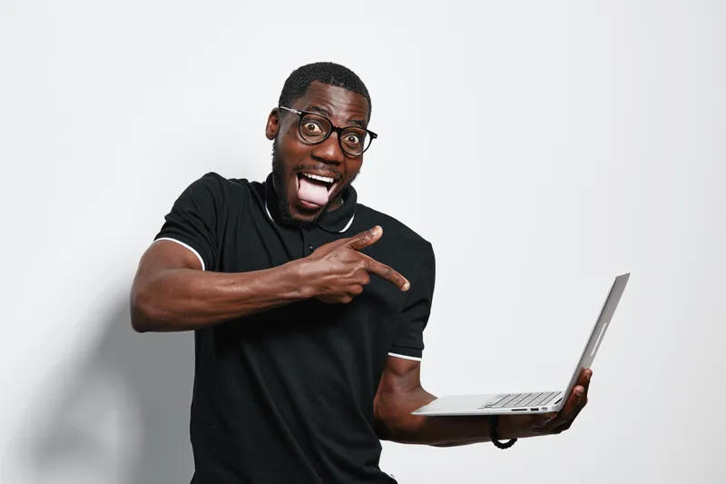African American man with dark skin and glasses holding laptop and pointing at screen with excited expression on white background in casual black shirt.