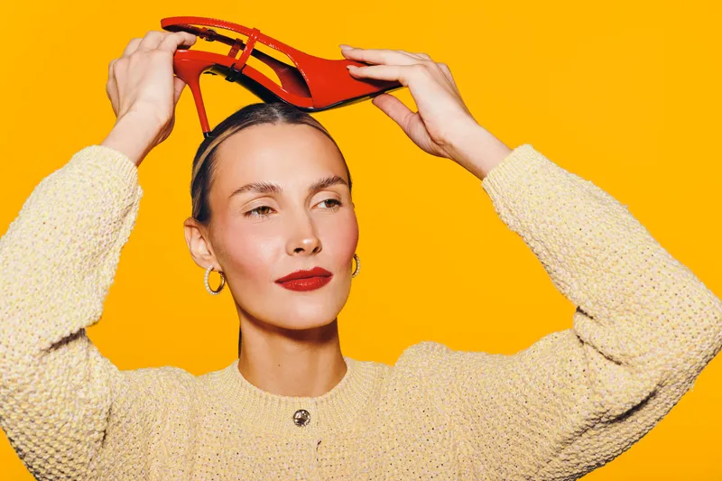 Young woman with red lipstick holds red high heel shoe above her head against bright yellow background. Fashion concept with stylish makeup and confident expression in studio lighting.
