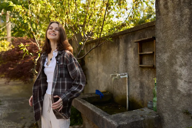 A young woman with plaid shirt smiles softly outdoors beside a stone wall, sunlit water trough creating a casual, carefree fashion moment