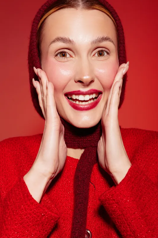 Happy woman in red sweater and hood smiles with hands on cheeks on red background. Concept of joy, fashion, winter style, and positive emotions in studio lighting.