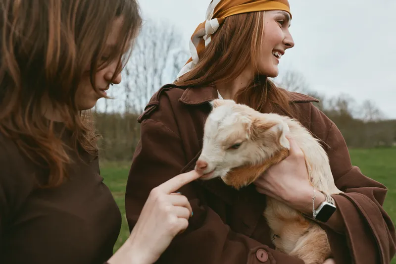 Lifestyle scene of two women outdoors with a gentle goat kid, showcasing affectionate interaction, joyful bond, warm light on a rural meadow, candid moment of care and friendship in natural colors,