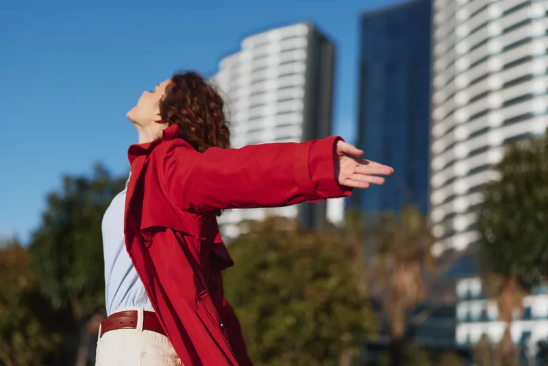 Young woman with curly hair wearing a red coat stands outdoors with arms outstretched enjoying fresh air and urban background on a sunny day.